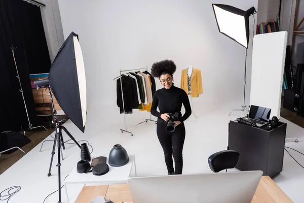 Productor de contenido afroamericano feliz con cámara digital cerca de ropa de moda y equipos de iluminación en el estudio de fotos - foto de stock