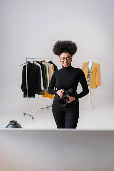 Produtor de conteúdo americano africano alegre com câmera digital perto da coleção de roupas da moda no estúdio de fotos — Fotografia de Stock