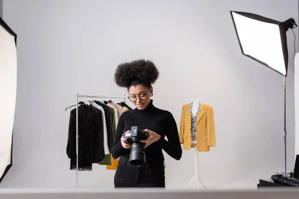 Pleased african american photographer looking at digital camera near fashionable clothes in photo studio — Stock Photo