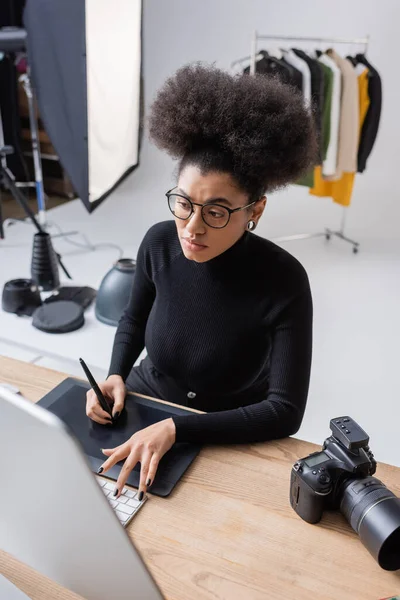 Visão de alto ângulo do retoque afro-americano em óculos trabalhando em tablet gráfico e computador perto de câmera digital em estúdio de fotografia — Fotografia de Stock
