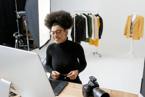 Feliz Africano americano fabricante de conteúdo olhando para monitor de computador perto de câmera digital e roupas da moda no fundo no estúdio de fotos — Fotografia de Stock