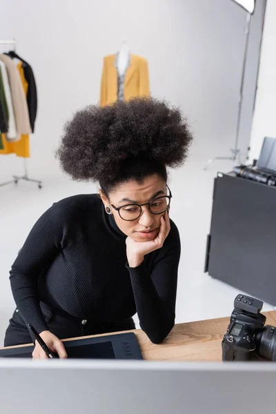 Retoque africano americano reflexivo en gafas mirando el monitor de la computadora mientras que trabaja en la tableta gráfica en el estudio de la foto - foto de stock