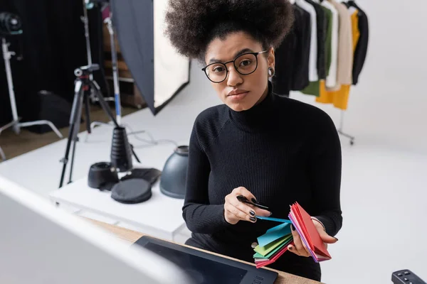 Ritocco africano americano in dolcevita nero e occhiali che tengono campioni di colore e guardano la fotocamera vicino al tablet grafico in studio fotografico — Foto stock
