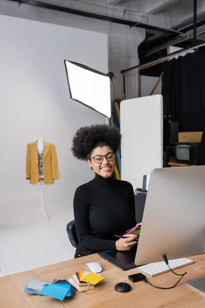 Alegre retoque afroamericano mirando a la cámara mientras se trabaja con muestras de color cerca de la tableta gráfica y el ordenador en el estudio de fotografía - foto de stock
