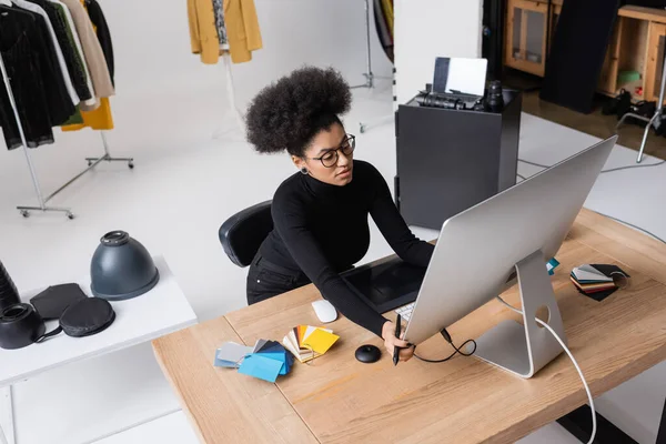 Vista de ángulo alto del retoucher afroamericano ajustando el monitor de la computadora cerca de la tableta gráfica y muestras de color en el escritorio en el estudio de fotos - foto de stock