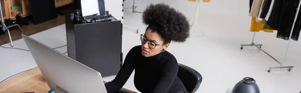 Vista de alto ángulo del gerente de contenido afroamericano en gafas y cuello alto negro trabajando en la computadora en el estudio de fotos, pancarta - foto de stock