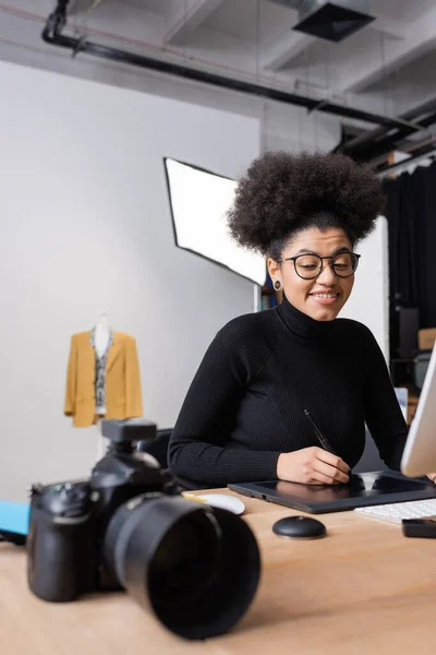 Alegre retoque afroamericano en gafas que trabajan en la tableta gráfica cerca de la cámara digital borrosa en el estudio de fotos - foto de stock