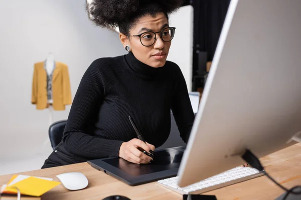 Retoque afroamericano concentrado en anteojos y cuello alto negro trabajando en tableta gráfica cerca del monitor de computadora en estudio fotográfico - foto de stock
