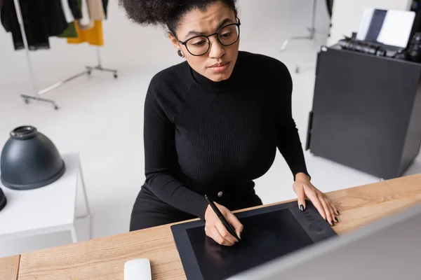 Retoque afroamericano enfocado en ropa negra y anteojos usando tableta gráfica mientras trabajaba en estudio fotográfico - foto de stock