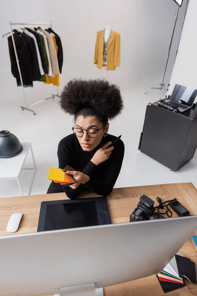 High angle view of african american retoucher looking at color swatches near graphic tablet and computer monitor in photo studio — Stock Photo