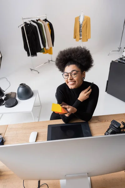 High angle view of cheerful african american retoucher holding color samples and looking at camera near graphic tablet in photo studio — Stock Photo