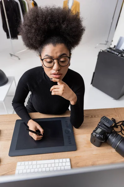 High angle view of thoughtful african american retoucher in eyeglasses working on graphic tablet and looking at monitor in photo studio — Stock Photo