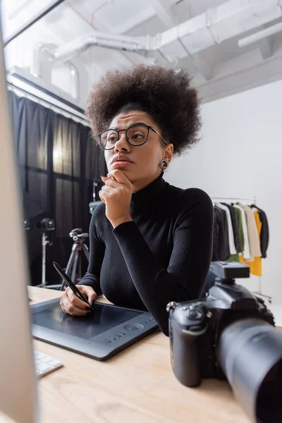 Pensive african american retoucher in black turtleneck and eyeglasses working on graphic tablet near blurred monitor in photo studio — Stock Photo