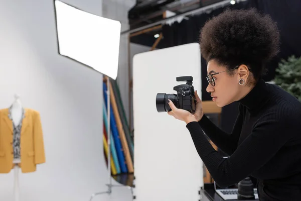 Vista lateral del fotógrafo afroamericano con cámara digital fotografiando chaqueta de moda en estudio fotográfico - foto de stock