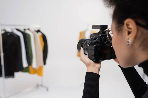 Photographe afro-américain avec appareil photo numérique prenant des photos de la collection de vêtements flous dans le studio photo — Photo de stock