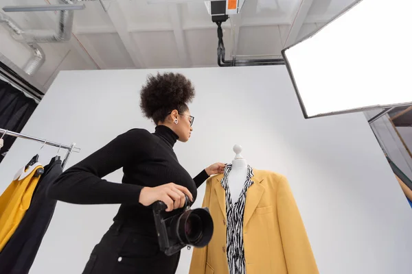 Vista a basso angolo del fotografo afroamericano con fotocamera digitale regolazione giacca elegante in studio fotografico — Foto stock