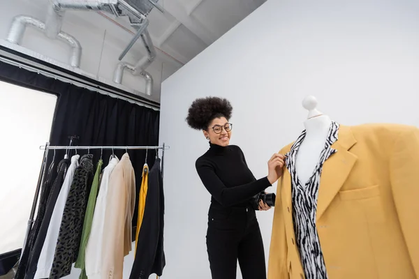 Joyeux photographe afro-américain regardant la caméra près des vêtements à la mode dans le studio de photo — Photo de stock