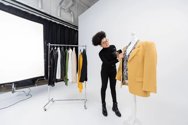Longitud completa de productor de contenido afroamericano con cámara digital ajustando chaqueta de moda en el maniquí en el estudio de fotos - foto de stock