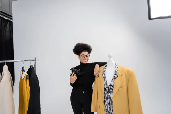 Alegre africano americano contenido fabricante con cámara digital posando cerca maniquí con chaqueta de moda en estudio de fotos - foto de stock