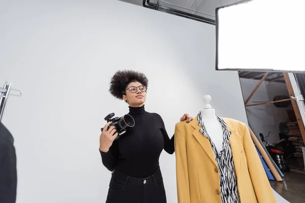 Fotógrafo afroamericano con cámara digital mirando hacia otro lado cerca maniquí con elegante chaqueta en el estudio de fotos - foto de stock