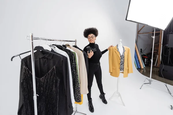Pleine longueur de fabricant de contenu afro-américain avec appareil photo numérique souriant près du support de rail et mannequin avec des vêtements à la mode dans le studio photo — Photo de stock