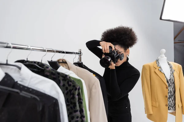 Fotógrafo afroamericano tomando foto de la colección de ropa de moda en el estante de ferrocarril en el estudio de fotos - foto de stock