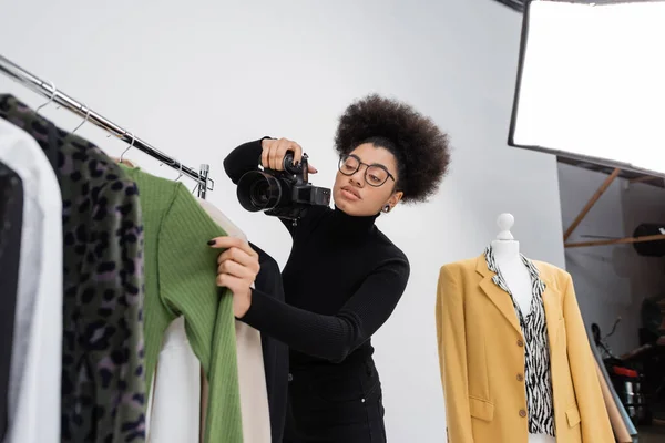Produtor de conteúdo americano africano em óculos tirando fotos de roupas da moda no estúdio de fotos — Fotografia de Stock