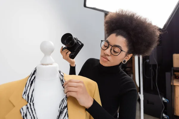 Productor de contenido afroamericano con cámara digital ajustando chaqueta de moda en maniquí en estudio de fotos - foto de stock