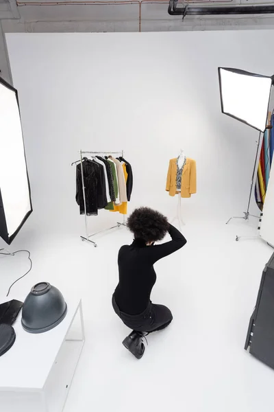 High angle view of african american photographer near rail rack with stylish clothes and lighting equipment in photo studio — Stock Photo
