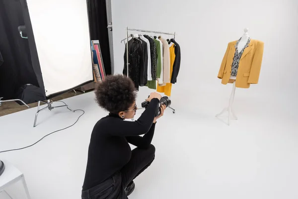 Fabricante de contenido afroamericano con cámara digital cerca de ropa de moda en rack de ferrocarril y maniquí en estudio de fotos - foto de stock