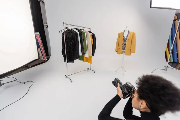 Fotógrafo afroamericano borroso tomando foto del estante de ferrocarril y maniquí con ropa elegante en el estudio de fotos - foto de stock