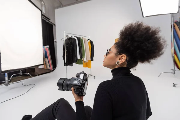 Fotógrafo afro-americano com câmera digital olhando para roupas na moda no estúdio de fotografia — Fotografia de Stock