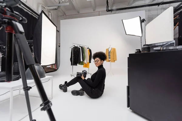 Piena lunghezza di allegro fotografo afroamericano guardando la fotocamera mentre seduto vicino alla collezione di vestiti alla moda in studio fotografico — Foto stock