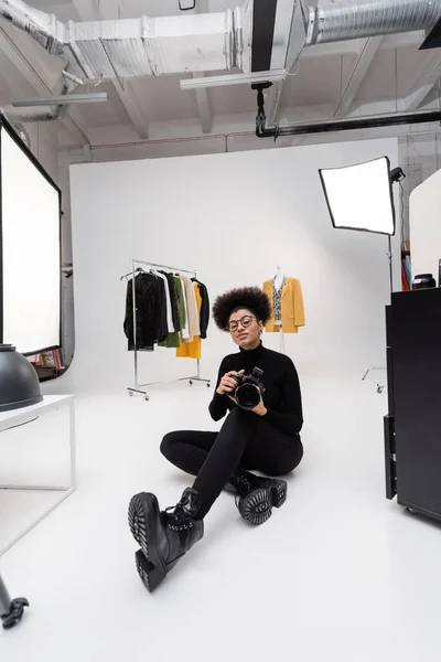 Full length of african american photographer in black stylish clothes sitting with digital camera in modern photo studio — Stock Photo