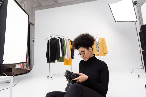 Fabricante de conteúdo afro-americano olhando para a câmera digital enquanto sentado perto de roupas da moda no estúdio de fotos — Fotografia de Stock