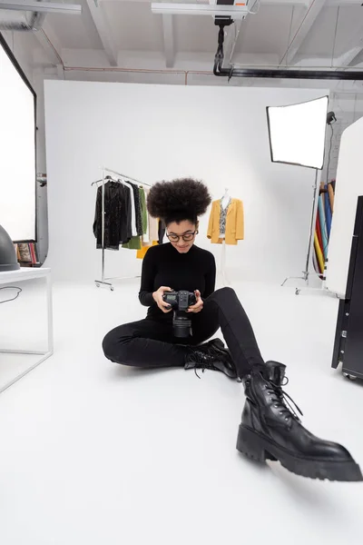 Piena lunghezza di felice produttore di contenuti africani americani seduto con fotocamera digitale vicino a faretti e vestiti alla moda in studio fotografico — Foto stock