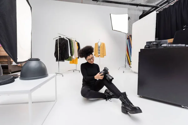 Photographe afro-américain souriant regardant l'appareil photo numérique tout en étant assis près des vêtements à la mode et projecteurs dans le studio de photo — Photo de stock