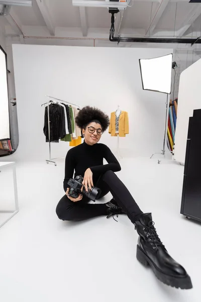 Produtor de conteúdo afro-americano em óculos e roupas pretas sorrindo para a câmera enquanto sentado no estúdio de fotografia — Fotografia de Stock