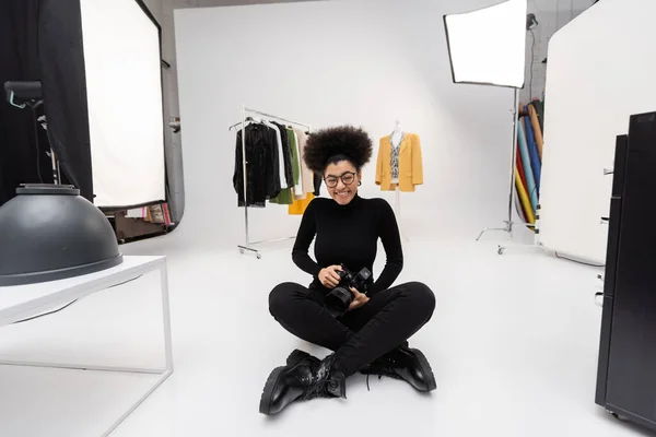 Alegre afroamericano fotógrafo con cámara digital sentado con las piernas cruzadas cerca de ropa de moda en estudio de fotos - foto de stock