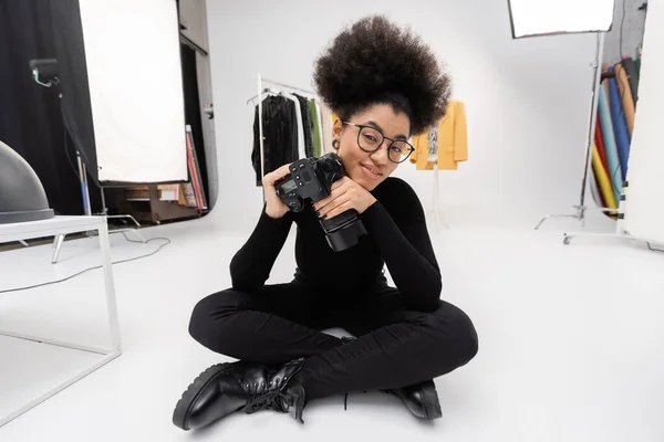 Femme afro-américaine souriante en vêtements noirs assis avec appareil photo numérique professionnel dans un studio photo moderne — Photo de stock