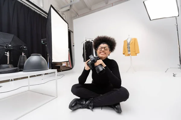 Productor de contenido afroamericano lleno de alegría con cámara digital sentada con los ojos cerrados en el moderno estudio de fotografía - foto de stock