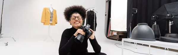 Радісний афроамериканський фотограф в окулярах посміхається закритими очима, сидячи в фотостудії, банер — стокове фото