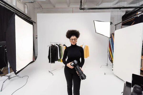 Mulher americana africana satisfeito com câmera digital em pé perto de holofotes e roupas da moda no estúdio de fotos — Fotografia de Stock