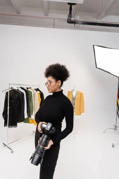Fotógrafo afroamericano en gafas y cuello alto negro de pie con cámara digital cerca de la colección de ropa de moda en el estudio de fotografía - foto de stock