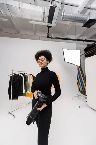 Positive african american photographer in eyeglasses standing with digital camera near rail rack with trendy clothes in photo studio — Stock Photo