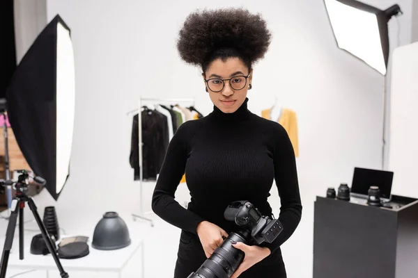Producteur de contenu afro-américain élégant en lunettes et col roulé noir debout avec appareil photo numérique en studio photo — Photo de stock