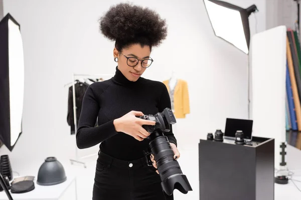 Fotógrafo americano africano em óculos e gola alta preta olhando para câmera digital profissional em estúdio de fotos — Fotografia de Stock