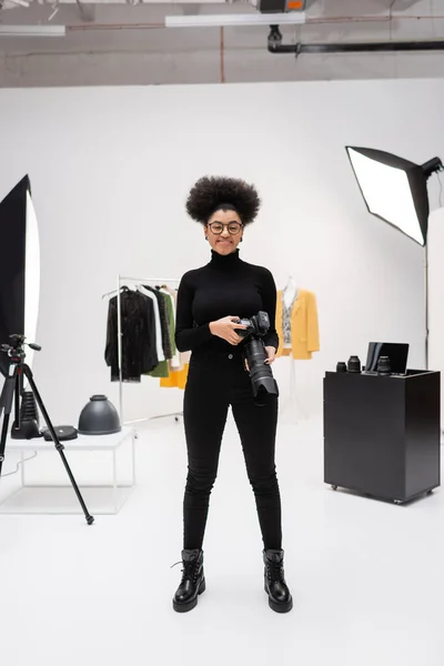 Longitud completa de fabricante de contenido afroamericano alegre en ropa negra de pie con cámara digital en estudio fotográfico moderno - foto de stock