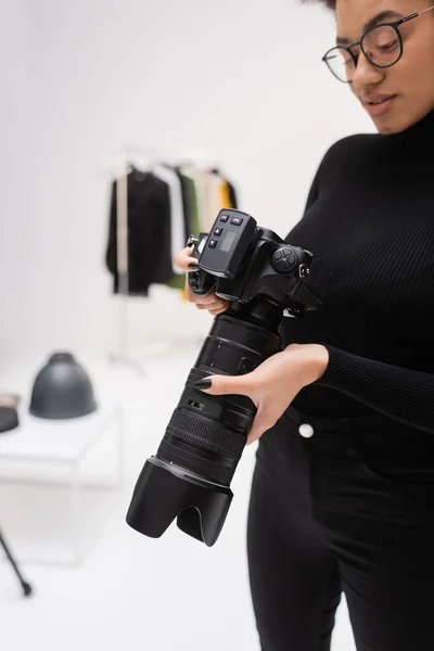 African american content maker in black turtleneck and eyeglasses holding professional digital camera in photo studio — Stock Photo