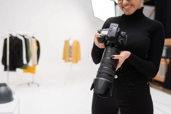 Vista parziale di allegro fotografo afroamericano in dolcevita nero con fotocamera digitale professionale in studio fotografico sfocato — Foto stock